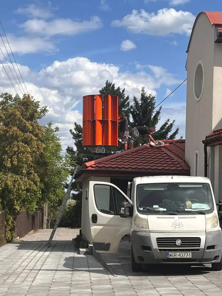 Dwóch pracowników montuje pomarańczową pionową turbinę wiatrową na dachu domu.